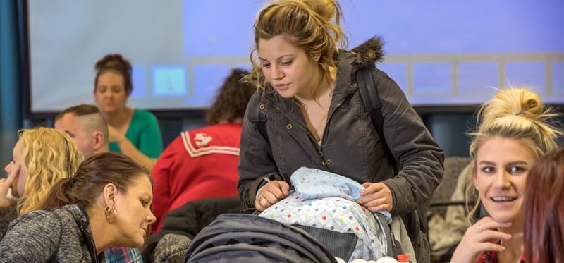 A young woman wheels her baby stroller through a group of people at a meeting.