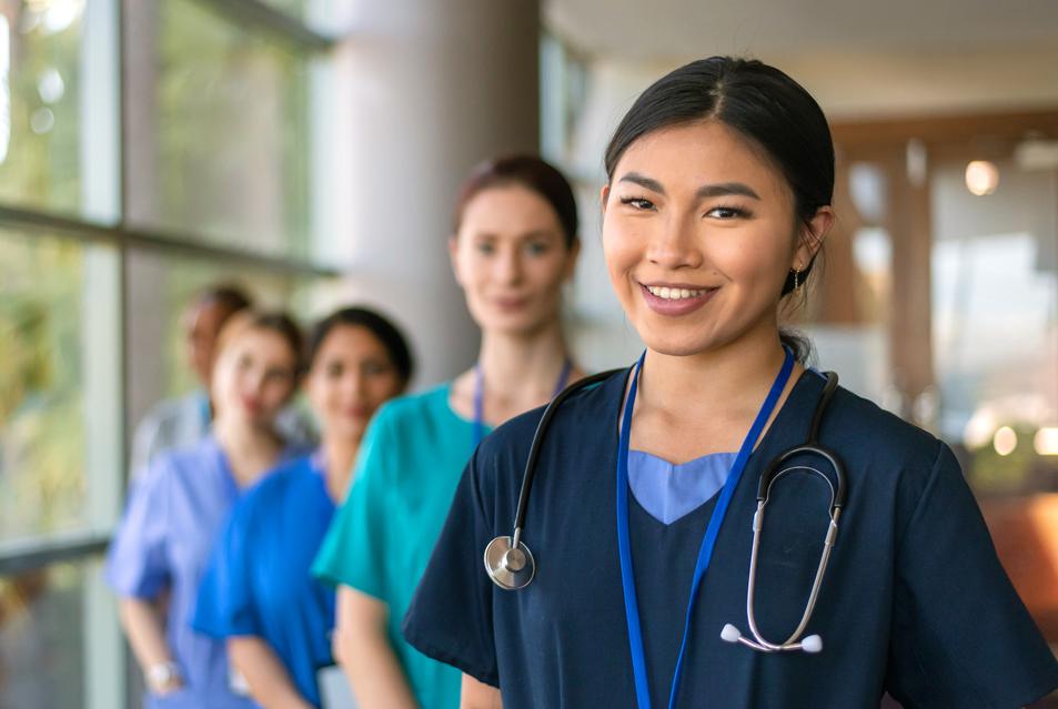 一名护士站在医院的大厅里，对镜头微笑，而其他一群护士站在她身后的其他护士则站着。