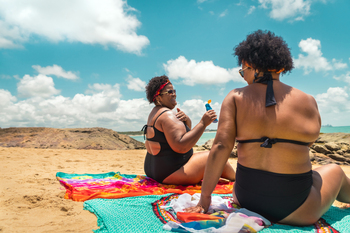 2 African American Women sunbathe on the beach on a bright summer day. One of the women is applying sunscreen.