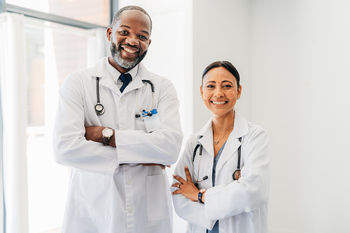 Two doctors stand in a hospital hallway with their arms crossed and smile for the camera.