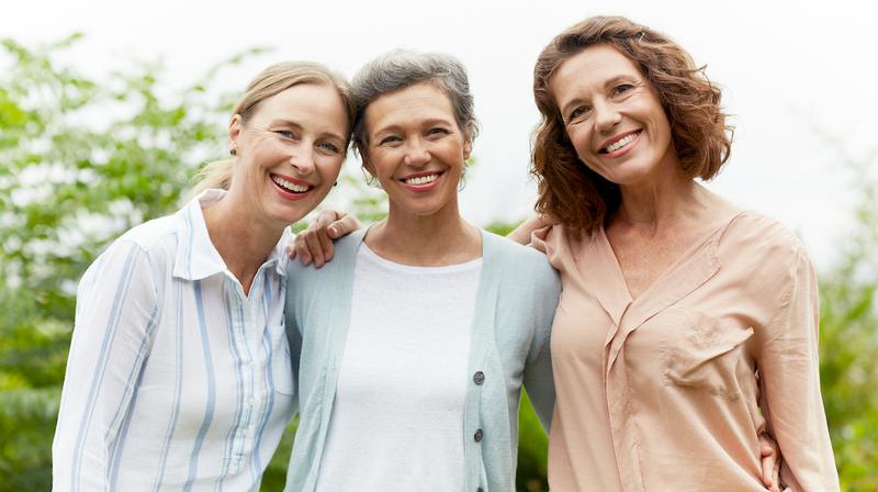 Three adult women friends smile for the camera with their arms around each others shoulders.