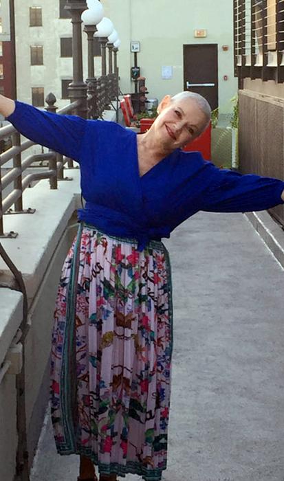 A woman with a shaved head and wearing a bright blue shirt and floral skirt smiles broadly as she stands on a balcony.