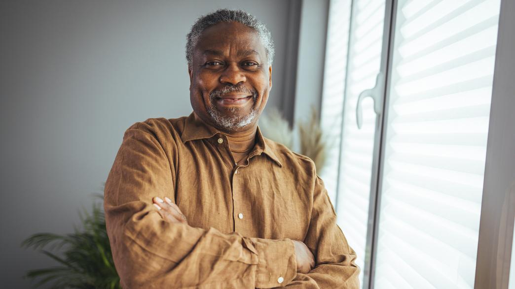 A mature african-american man stands next to a window in a home with his arms folder and smiles for the camera.