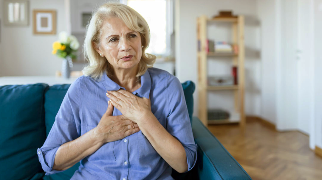 A woman sits on her livingroom sofa and puts her hands on her chest indicating she is in pain.