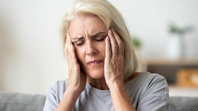 A middle aged woman winces in pain as she holds her hands to her temples. She is suffering from a migraine headache.