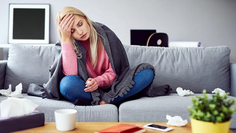 A woman sits on her sofa under a blanket while sick at home.