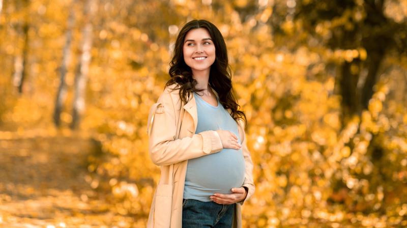 A pregnant woman poses for a photo with her hands on her belly outdoors with fall foliage in the background.