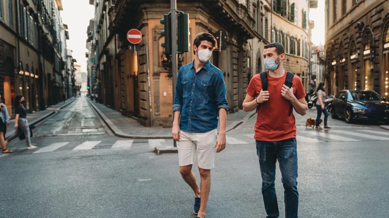 Two young men walk and talk together in an urban environment while wearing masks.