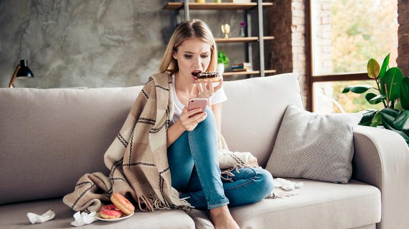 A young woman eats a snack while sitting on the couch in her living room and looking at her phone.