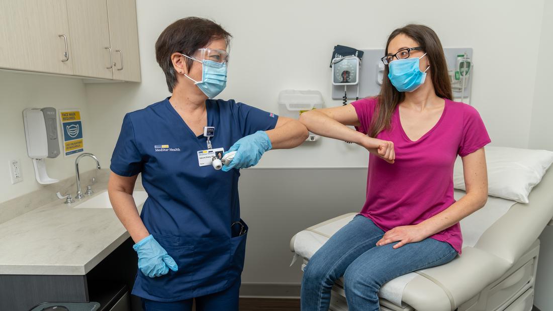 A healthcare worker bumps elbows with a patient in a clinical setting. Both people are wearing masks.