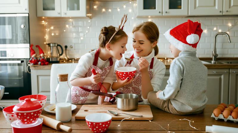 A mother and her 2 young children cook together in the kitchen at holiday time. Kids are wearing reindeer antlers and a santa hat.