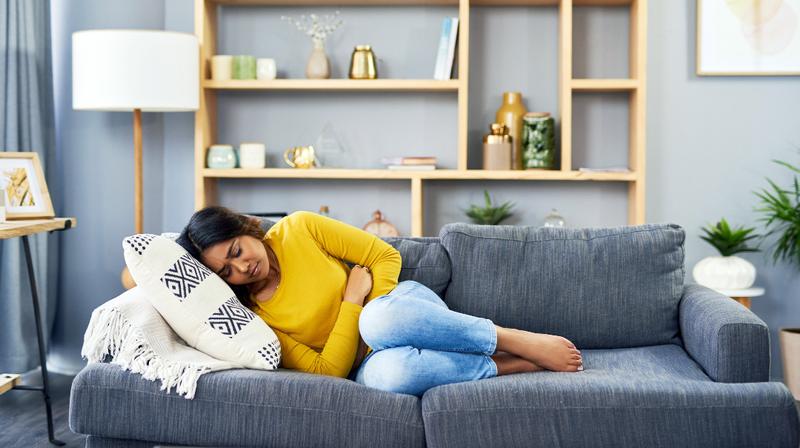 A young woman lays on the couch at home, holding her stomach and wincing in pain.