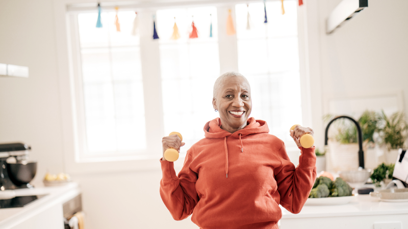 A senior woman smiles as she exercises at home with hand weights.