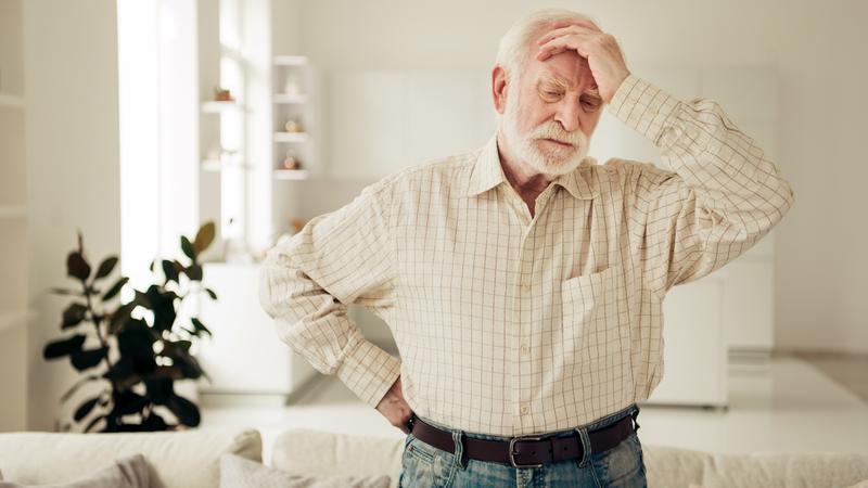 A senior man holds his hand to his head while standing in his livingroom at home.