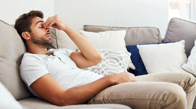 A man holds the bridge of his nose in pain while laying on a sofa at home.