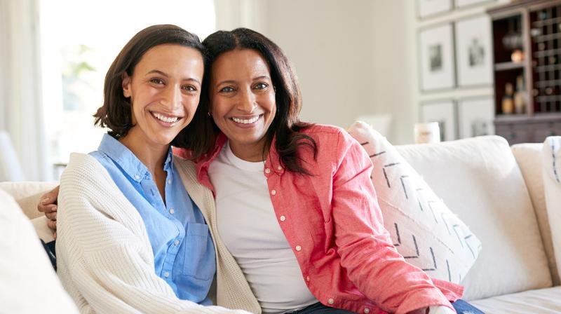 Two women sitting close together on a sofa in a white living room and pose for a photo.