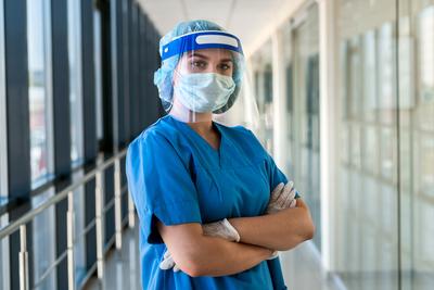 A healthcare provider, dressed in full PPE gear, stands with her arms crossed and looks at the camera in a hospital hallway.