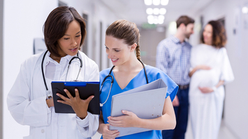 Two medical professionals reviewing information in a hospital hallway with other patients in the background.