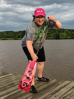 辛迪·戴维斯站在湖的码头上，手里拿着粉红色的桨。辛迪是一位乳腺癌幸存者，她过着积极的生活方式。