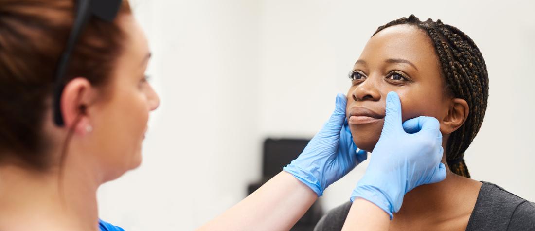 A female doctor wearing blue gloves examines the face of a female patient.