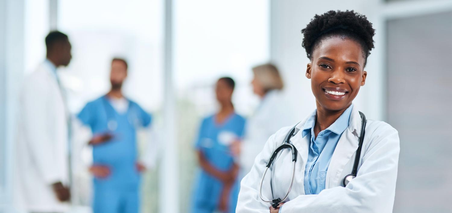 A female African American doctor stands with her arms crossed, stethescope around her neck and smiles as she looks into the camera.