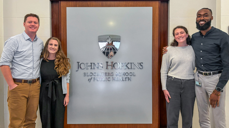 A group of 4 residents stand in front of the Johns Hopkins Bloomberg School of Public Health entrance.