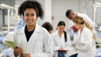 A female medical student holding a notebook smiles for the camera. A group of students are blurred in the background.