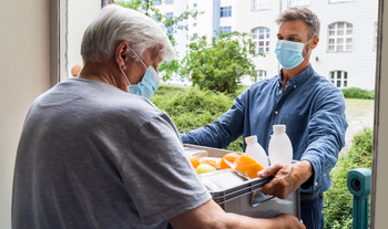 An elderly man receives a delivery of food at his home. The man and the delivery person are both wearing masks.