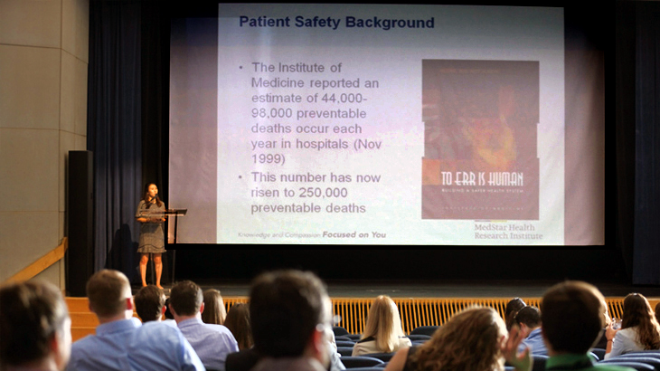 A woman speaks behind a podium while a large screen displays information during her presentation in a large auditorium at a medical conference