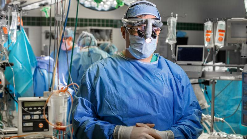 Dr Ezekiel Molina wears full operating protection and stands for a portrait in an operating room at MedStar Washington Hospital center. He is wearing a mask, gown, face shield, a surgical skull cap and magnifying headset.