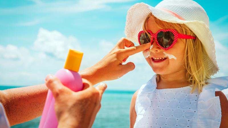 Close up photo of a little girl's face, wearing heart sunglasses, as her mother applies sunscreen lotion to her daughter's face.