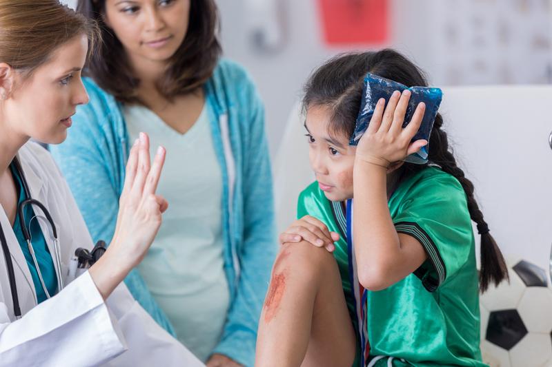 A doctor holds up 3 fingers in order to test a young pediatric patient for concussion. The girl has an ice pack on her head.