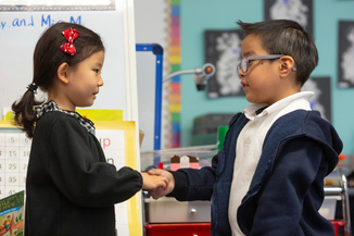 A young girl and a young boy shake hands in a school classroom.