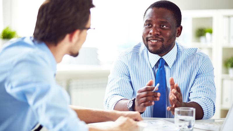 Two professional men sit at at table and talk.