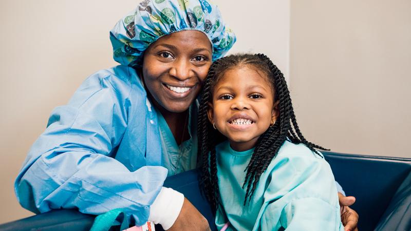 A dentist wearing blue scrubs and a blue hair covering, poses with a young girl patient.