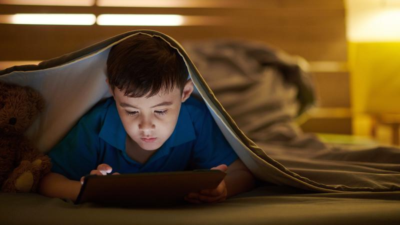 A young boy looks at an ipad while under the covers in his bed.