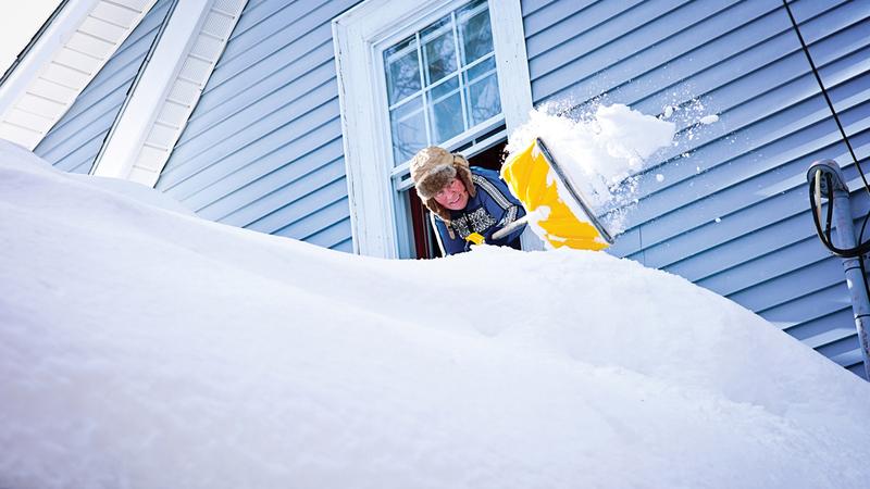 A man shovels a large pile of snow while leaning out of a window in his house.