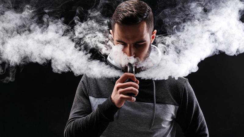 A man uses a vaping device. It is dramatically lit to highlight large plumes of vapor.