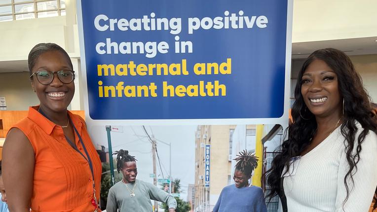 Roxane Richardson and Jodi Ann Boreman stand together at a conference next to a banner for the Safe Babies Safe Moms program.