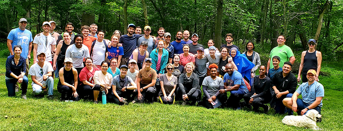 A large group of MedStar Health graduate medical education residents sit together for a photo outside in a park.
