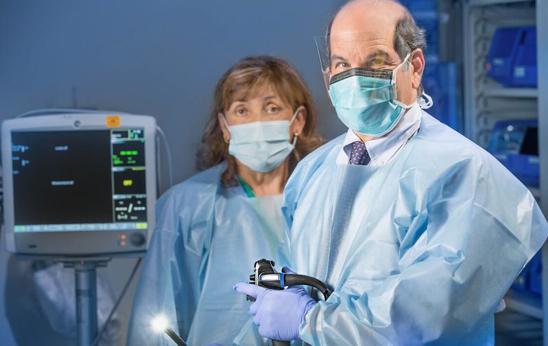Dr David Shocket holds a colonoscope in a treatment room at MedStar Health. A female physician assistant stands behind him. Both people are wearing blue scrubs and masks.
