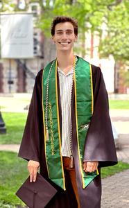 扎克·斯坦戈，MMMC女性董事会奖学金获得者，在校园树荫下合影留念。他穿着一件黑色的毕业礼服，配着绿色的领口饰带，戴着一顶毕业帽。