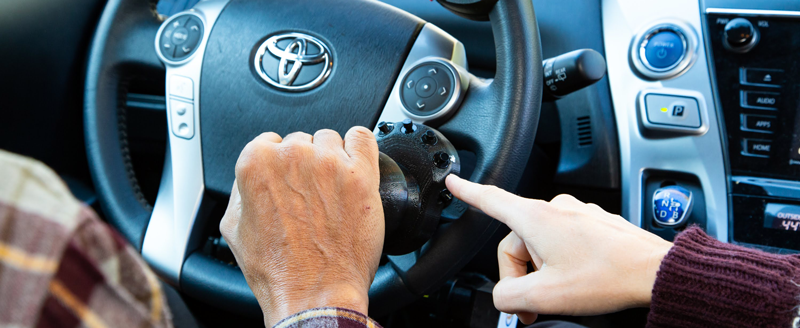 Close up photo of two people's hands on a car steering wheel.