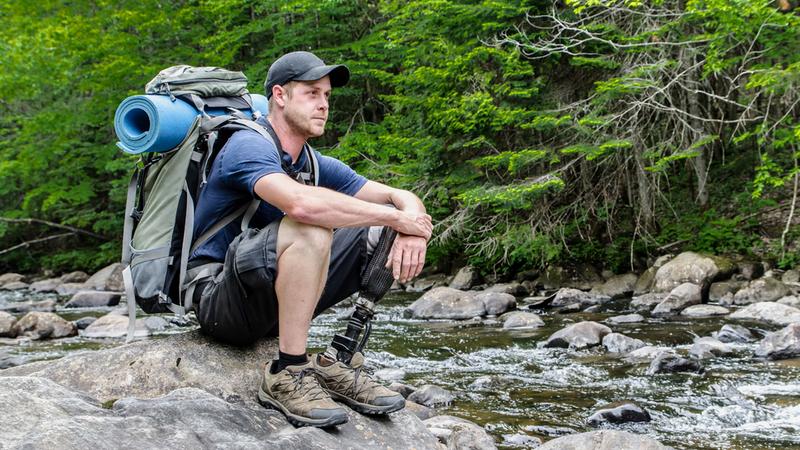 A man with a prosthetic leg sits on a rock at the shore of a river to take a break while hiking.
