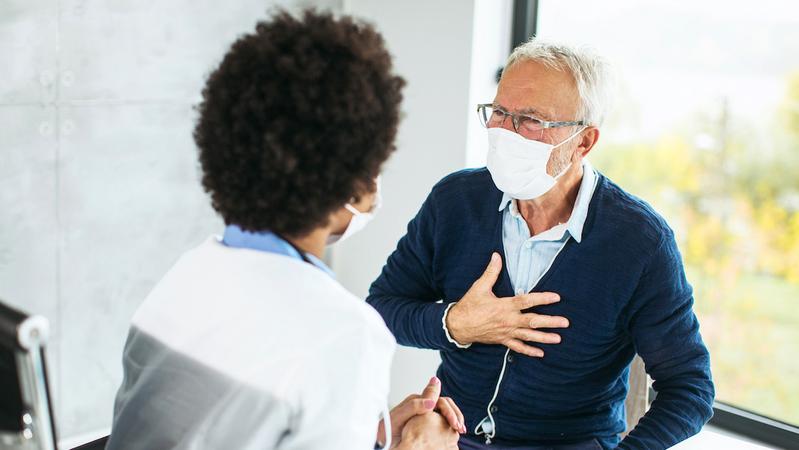 A senior male patient talks with his doctor in a clincial setting. Both people are wearing masks.