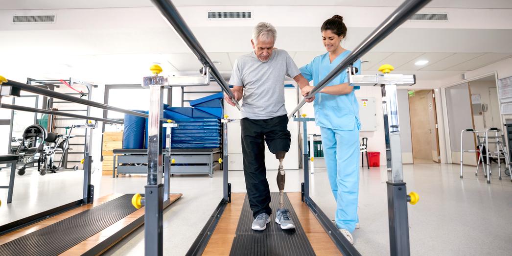 A senior man wearing a prosthetic leg works with a physical therapist at a rehabilitation gym.