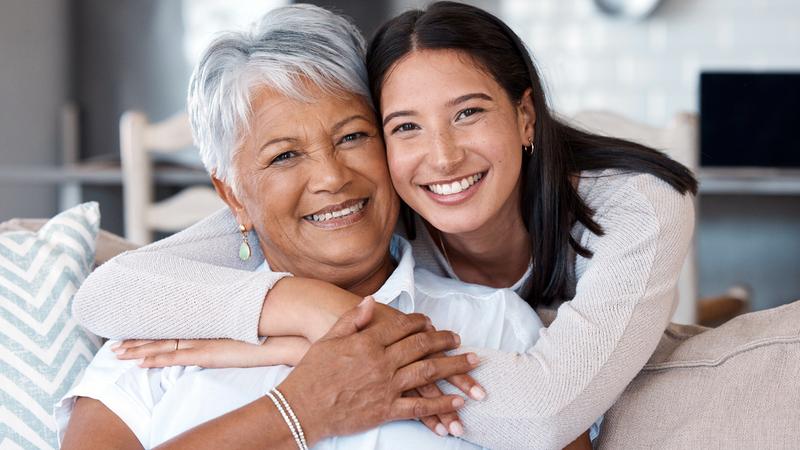 A young woman hugs her mother while she sits on a sofa in her home.