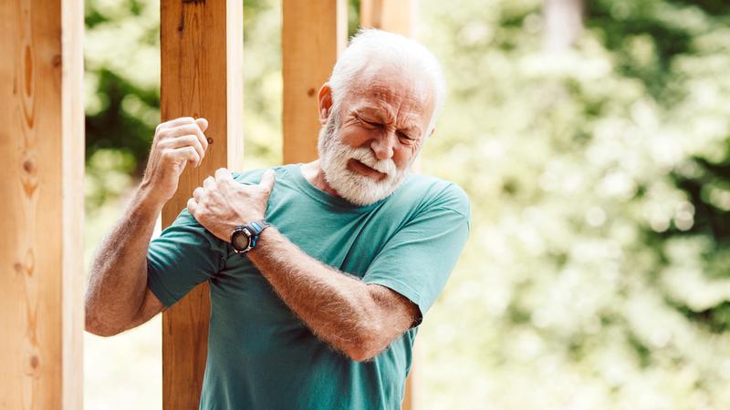 A senior man holds his shoulder as he winces in pain while standing near a pull-up bar at an outdoor fitness station in a park.