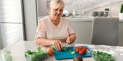 A woman sits at a table in her kitchen and cuts vegetables for a salad.
