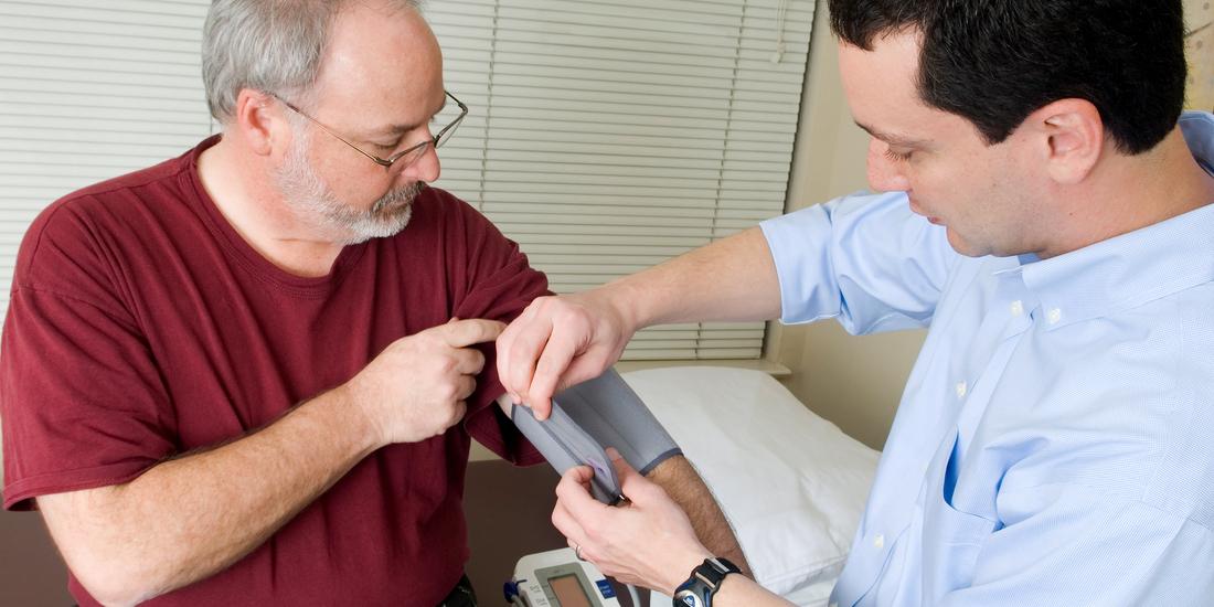 A doctor takes the blood pressure reading for an senior male patient in a clinical setting. The photo is taken from a high camera angle.
